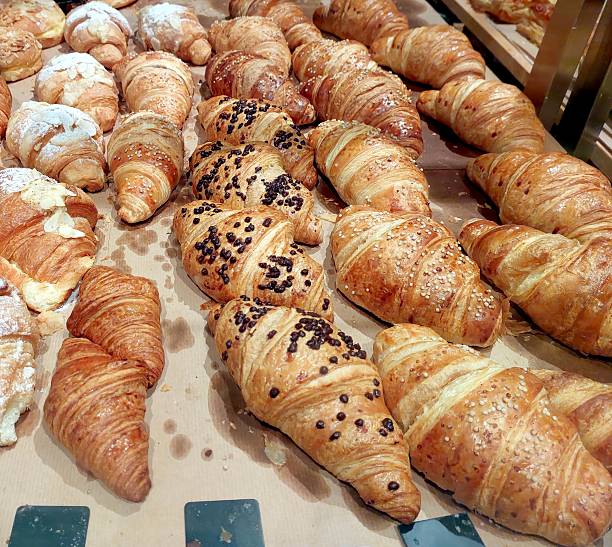 Croissants of various styles and toppings sit on a baking sheet, freshly baked and ready to be enjoyed in a welcoming bakery environment.