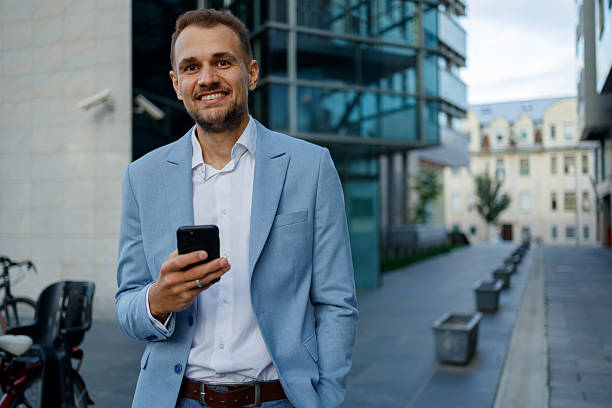 istock Young businessman smiling, using smartphone outdoors in urban city setting, looking at camera 2236334211