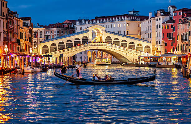 Evening view of gondolas on the Grand Canal with the historic Rialto Bridge lit up in Venice, Italy.