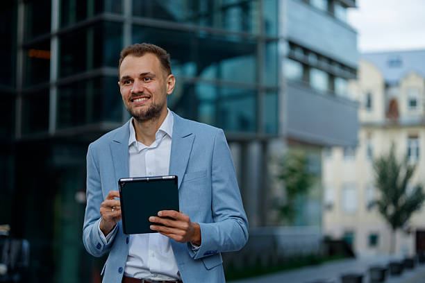 istock Smiling businessman using a digital tablet while walking in a modern city street, managing business remotely 2236334270