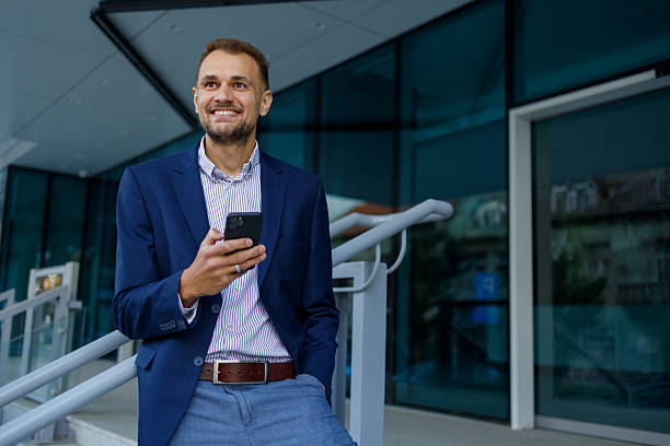 istock Businessman holding mobile phone, smiling while looking away, standing outside modern office building 2236334685