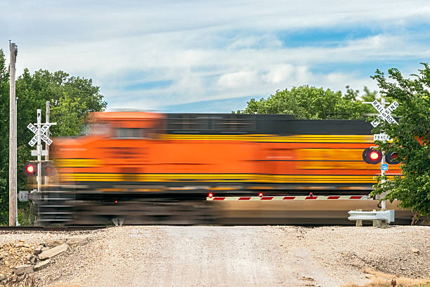 Dangerous rural railroad crossing as bright orange locomotive passes at speed: dirt road with steep approach; tree-lined; boom barrier with flashing lights active; railway warning signs and red stop lights active; in-camera motion blur. ID concealed in blur.