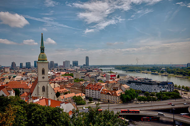 istock Panoramic view over Bratislava from the castle hill, with St. Martin’s Cathedral in the foreground, the Danube River winding through the city, and modern high-rises forming the skyline under a bright summer sky. 2236357345
