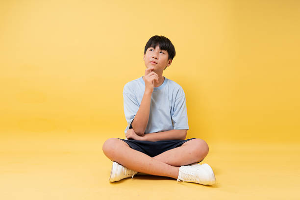 istock Boy in blue shirt sitting cross-legged with thoughtful expression on a yellow background 2236369376