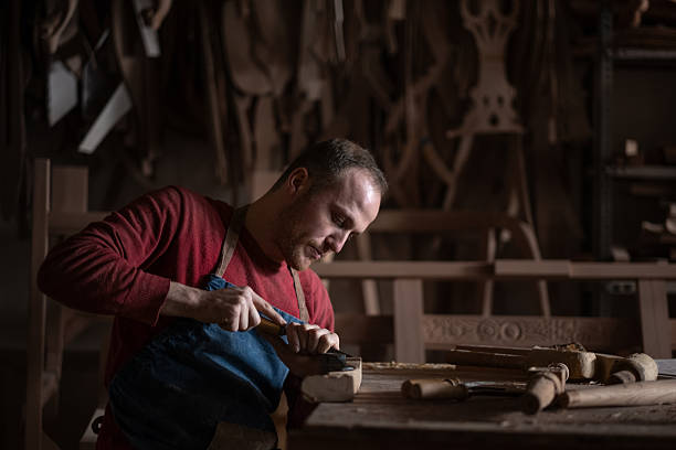 Male artisan concentrating, skillfully carving wood with hand tools in a dim traditional workshop
