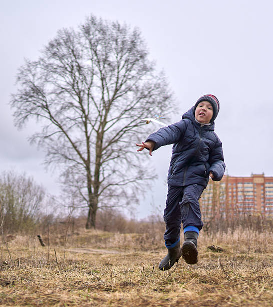 A cheerful elementary age boy dressed in casual outerwear and rubber boots is holding the handmade toy airplane, playing with in and throwing it. Shooting at overcast spring day in a countryside near town