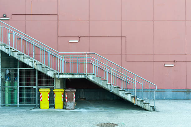 The exterior of a modern building with a façade characterized by rectangular panels. A concrete staircase with a metal railing ascends diagonally. Three garbage cans are positioned beneath the staircase. The scene depicts an urban environment.