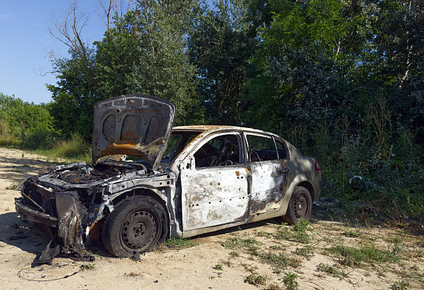 istock A heavily burned-out car with its hood open sits abandoned on a dirt path under a clear sky, surrounded by green trees and bushes. 2236387553