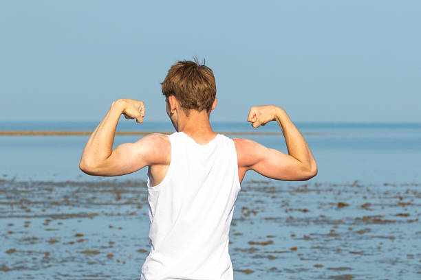 Teenage boy flexing his bicep arm muscles wearing a sleevless top and shorts on a beach from behind