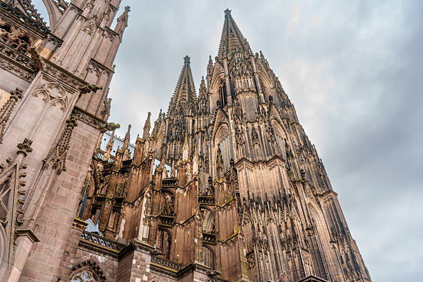 Cologne, Germany - June 15, 2025: Detailed view of the intricate Gothic spires and facade of Cologne Cathedral, featuring ornate stonework, towering structures, and a moody sky.