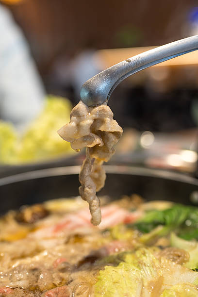 Hands holding chopsticks dipping a slice of pork into a bubbling shabu shabu pot filled with flavorful broth.