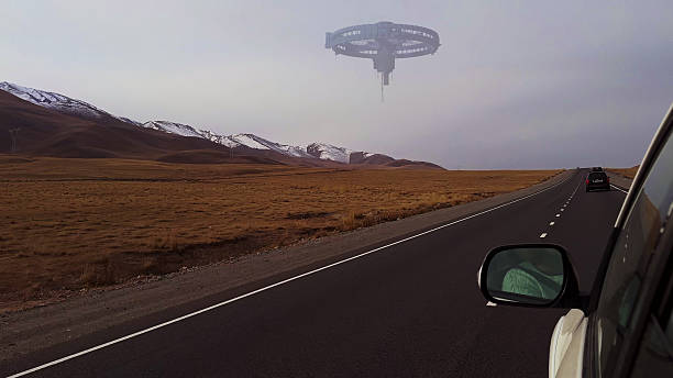 An alien spacecraft hovers in the distance above a straight road stretching through barren mountain terrain.
