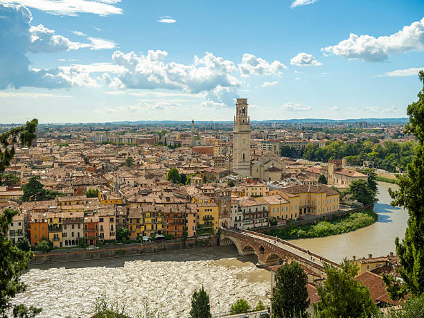 A panoramic view of Verona, Italy, showcases the Adige River, historic Ponte Pietra bridge, and Lamberti Tower under a dramatic sky. The vibrant old town glows with warm colors and timeless charm.