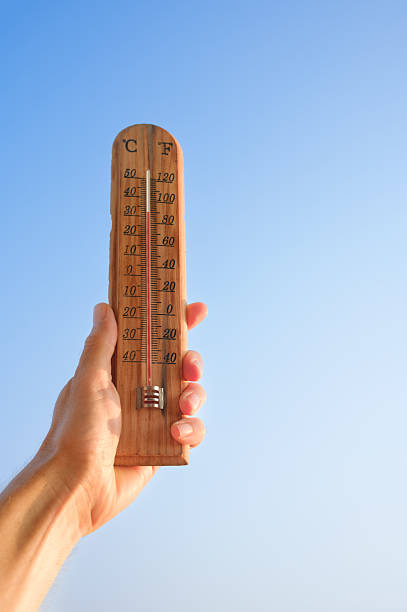 Hand holding a wooden thermometer against a clear blue sky, showing the mercury rising to high temperatures, representing extreme heat, global warming, and climate change concepts