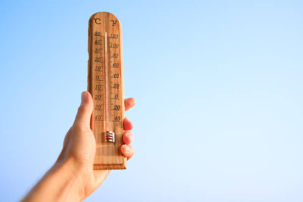 Hand holding a wooden thermometer indicating a high temperature, symbolizing summer heat, global warming, and extreme climate conditions with a clear blue sky in the background