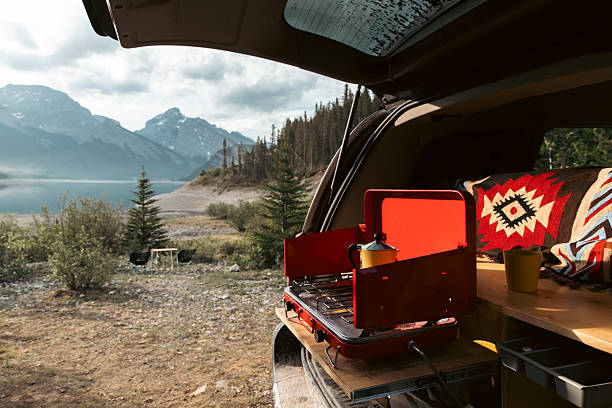 A camper’s mobile kitchen inside the open trunk, featuring a red stove, kettle, and a woven blanket. Mountain lake scenery, pine trees, and distant peaks convey a peaceful, adventurous outdoor retreat.