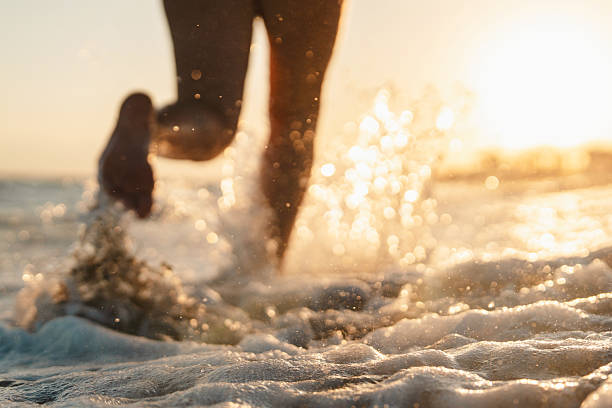 Woman splashing waves while running on the beach at sunset, unrecognizable person, low section
