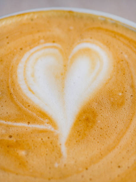 A beautifully crafted cup of coffee featuring a heart design sits on a white saucer in a charming cafe. Natural light enhances the warm tones, creating an inviting atmosphere for relaxation.