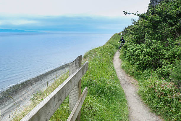 A person riding a mountain bike rounds a corner on the Bluff trail at Fort Ebey State Park.  The Straits of Juan de Fuca is in the background.  A fence borders the path.