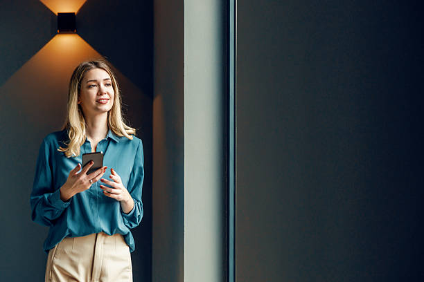 istock Smiling woman in a blue blouse holding a smartphone while standing by a large window in a modern indoor space 2236452487