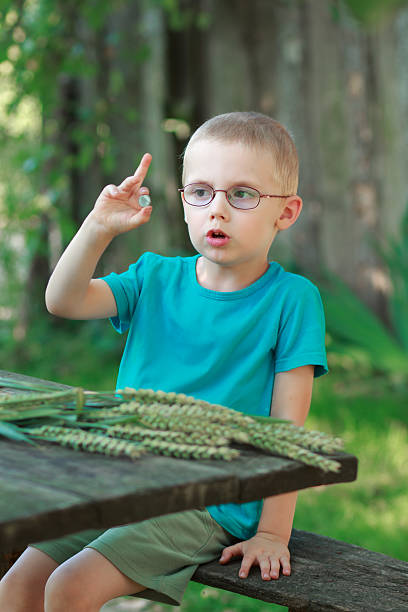 A boy in glasses sits at a wooden table outdoors, holding a coin and talking with an expressive gesture. Summer holidays, healthy outdoor activities.