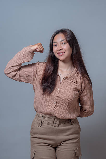 istock Confident Indonesian woman smiling while flexing her arm and showing off her strength in a beige blouse and pants. The photo captures an expression of empowerment against a neutral gray background. 2236468230