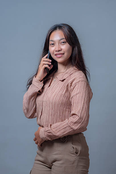 istock Confident young Indonesian woman smiling while talking on a smartphone. She stands against a neutral background, portraying modern communication, digital lifestyle, and smart technology use. 2236469355