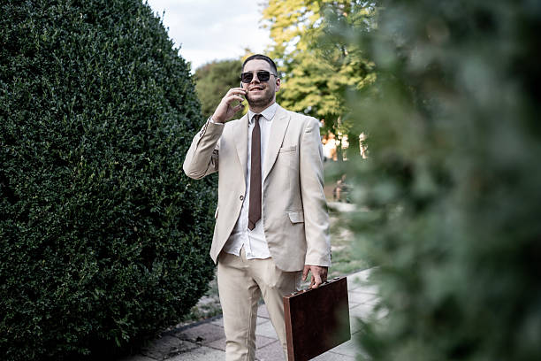 A stylish businessman wearing a light beige suit and sunglasses stands on a sunny park path, speaking on a mobile phone while holding a brown briefcase, exuding confident urban professionalism.