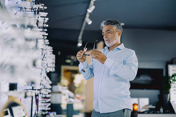 istock Senior man browsing eyeglass frames in a modern optical store. He is standing near a display wall, thoughtfully considering his options. The store has a clean, bright interior with a wide variety of eyewear on display. 2236475458