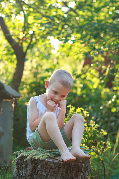 A barefoot boy sits on a stump with wheat stalks, holding his fists to his chin in a shy pose. Summer holidays, outdoor recreation for children.