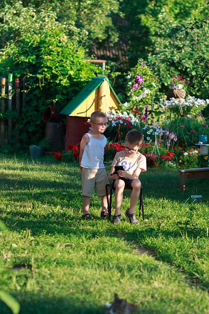 Brothers in glasses spending time outdoors, one focused on smartphone while the other stands nearby. Childhood and technology concept.