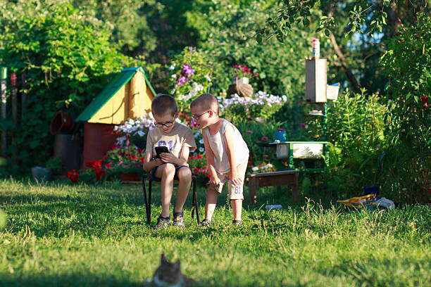 Two kids on summer holidays sitting in the garden, watching smartphone together. Concept of unity and togetherness.