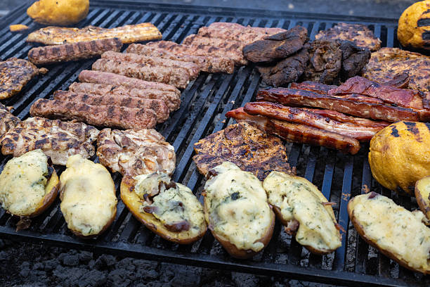 variety of grilled meats and roasted potatoes on a barbecue. The image captures the textures and colors of the food, including grilled sausages, pork ribs, and marinated chicken, alongside cheesy roasted potatoes