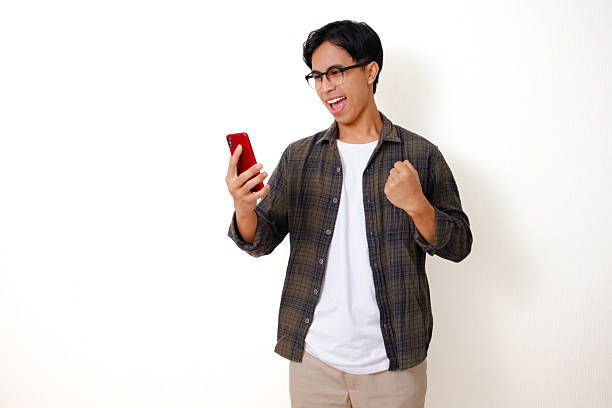 istock Excited young Asian man celebrating success while looking at his mobile phone and clenching his fist, isolated on white background 2236500083
