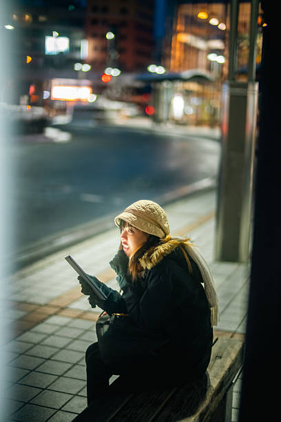 Asian woman remote working freelance on digital tablet while waiting for bus in winter night. Woman traveler enjoy urban lifestyle travel in the city with using wireless technology and gadget device.