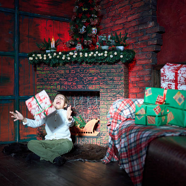 A cheerful elementary age boy dressed in casual clothes is waiting Christmas. He is sitting by a fake fireplace with a gift in his hands. The traditional red and green colors in Christmas decor of the living room. Studio shooting