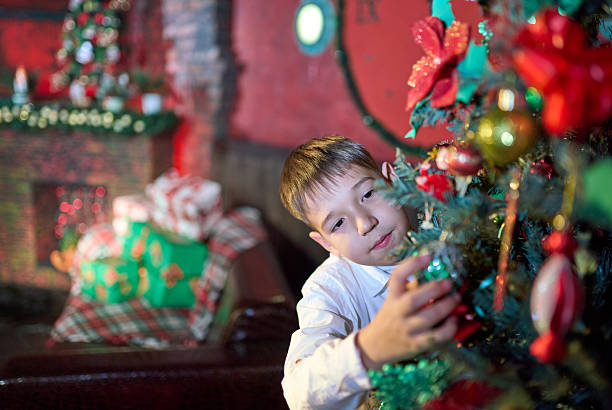 A cheerful elementary age boy dressed in casual clothes is waiting Christmas. He looks out from behind the tree, looking at the Christmas tree decorations on it. The traditional red and green colors in Christmas decor of the living room. Studio shooting