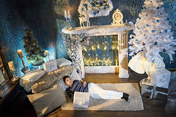 A cute elementary age boy is spends time waiting for Christmas at a room decorated at Christmas. He is standing, holding the gift box, smiling and dreaming with closed eyes. Studio shooting