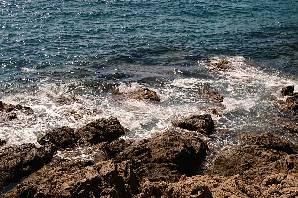 Sunlight reflecting on turquoise sea waves hitting rugged coastal rocks. Tranquil Mediterranean seascape with natural textures and vivid contrast between water and stone - captured in warm daylight, evoking calm and freedom.