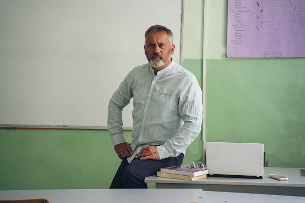 Mature male teacher or professor in a classroom, using a smartphone and a laptop. Scenes show him thinking, writing, teaching, and engaging with modern technology. Surrounded by desks, a whiteboard, and educational materials. Represents a contemporary educator in a professional setting. Ideal for themes of education, digital literacy, learning, and teaching.