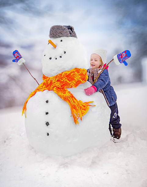 Smiling little girl hugging a big snowman decorated with a carrot nose, fur hat, orange scarf, and mittens in a snowy winter landscape. Perfect for Christmas, childhood, family fun, and seasonal holidays.
