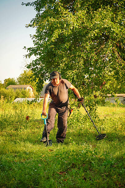 istock A man uses a metal detector in a green field, focused on finding hidden objects in the grass under the warm light of late afternoon. The area is surrounded by trees and distant structures 2236550710