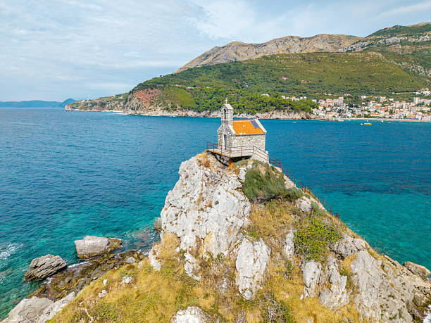 Small stone chapel perched on rocky cliff overlooking clear blue sea. Scenic coastal landscape with hillside town in background. Remote religious building by the ocean.