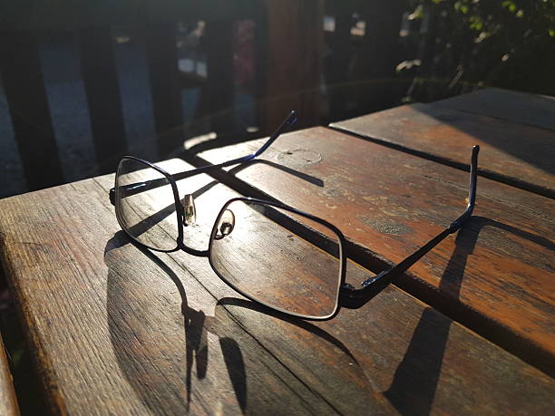 A pair of eyeglasses lies on a wooden table, casting a distinct shadow in the warm glow of the setting sun