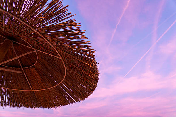 Close-up of a straw beach umbrella silhouetted against a vibrant sunset sky. The intricate structure of the umbrella’s edge is in sharp focus, while the sandy beach, gentle waves, and pastel-toned sky fade softly into the background. The warm hues of orange, pink, and lavender create a tranquil and dreamy seaside atmosphere.