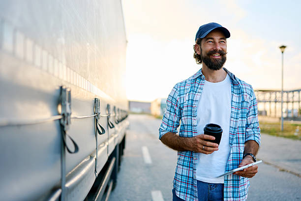 Smiling truck driver stands beside a semi trailer, holding coffee and a tablet during a daytime break, checking route and planning next delivery with relaxed confidence