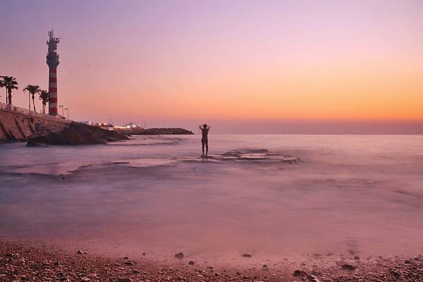 Long-exposure seascape at sunrise along the Mediterranean coast in Lebanon, featuring a striped lighthouse and rocky shoreline. The soft pastel sky blends orange, purple, and pink hues, reflecting over the calm sea waves. A symbolic view of tranquility, coastal heritage, and the beauty of Lebanon’s maritime landmarks.
