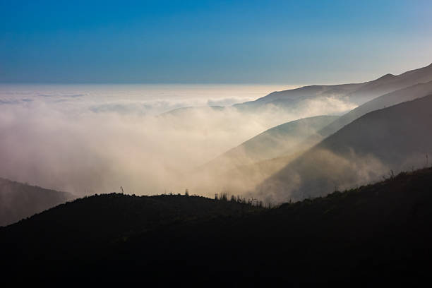 A wide-angle, high-angle view captures the scenic Pacific Palisades coastline from a dry hillside trail, with a low-lying marine layer of fog rolling over the Pacific Ocean below a clear blue sky in September.