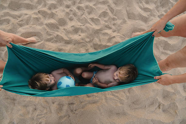 Two young boys relaxing in a makeshift hammock on a sandy beach, held by adults