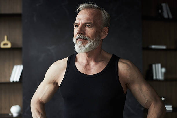 Middle aged Caucasian man standing with hands on hips, looking away from camera, showing muscular arms and confident posture, short gray hair and full beard visible